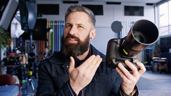 Bearded photographer holding a telephoto lens in a studio setting, demonstrating camera equipment.
