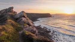 Rocky coastal cliffs overlooking turbulent ocean waters at golden hour sunrise or sunset.