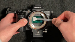 Hands demonstrating sensor cleaning on a Sony Alpha camera with visible mirror and sensor elements.