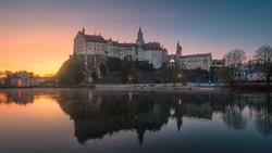 Medieval castle with multiple spires reflected in still river water at sunrise.