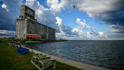 Abandoned grain elevator tower beside a calm waterfront with Adirondack chairs on the grass.