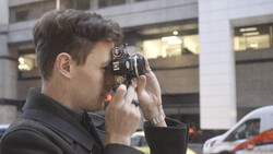 Photographer holding a vintage film camera up to his face to compose a shot outdoors.