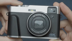 Compact digital camera with black body and silver top plate held in hands against gray background.