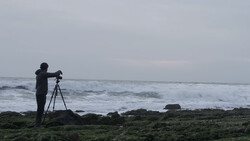 Photographer on tripod positioning equipment on rocky shore facing turbulent ocean waves.