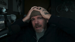Man with beard and tattoos shading his eyes while speaking into a studio microphone.