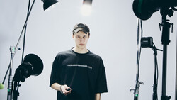 Man in black t-shirt and cap stands in professional studio surrounded by lighting equipment.
