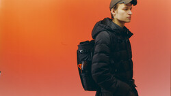 Young man in black puffer jacket and cap standing against warm orange gradient background.