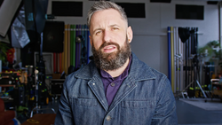 Bearded man with gray hair wearing denim jacket over purple shirt in workshop studio.