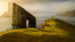 Moss-covered coastal cliff with natural rock arch framing calm sea and distant headlands.