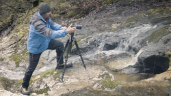 Photographer on tripod shooting a flowing stream in a moss-covered woodland setting.