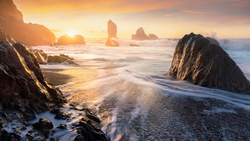 Rocky sea stacks silhouetted against golden sunrise over a misty ocean beach.