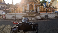 Camera on tripod with articulating monitor facing an ornate Buddhist temple with golden spires and white walls.
