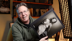 Photographer holding a black and white portrait book in a studio setting with bookshelves behind.