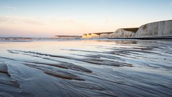 White chalk cliffs reflecting in wet sand at golden hour, with calm sea and distant headland.