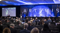 Speaker presenting at a conference with large blue screen displaying ARTEMIS backdrop.