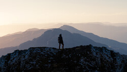 Silhouetted hiker standing on a rocky outcrop overlooking layered mountain ranges at sunset.