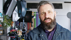 Bearded man with gray hair in denim jacket poses in front of photography studio setup with lighting equipment and shelves of gear.