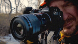 Photographer holding Olympus camera with telephoto lens against bare trees in outdoor setting.