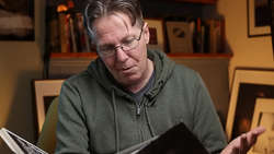 Man wearing glasses and olive green hoodie reviewing printed photographs at a table.