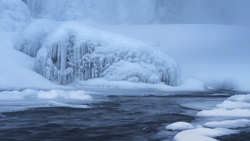 Frozen waterfall and icicles along an unfrozen river channel surrounded by snow and ice.