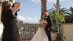 Wedding photographer capturing bride and groom posing on a terrace with palm trees and ocean view.