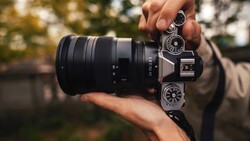 Photographer holding a mirrorless camera with attached telephoto lens, displaying the camera's top controls and dial interface.