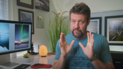 Man in teal shirt gesturing while discussing photography at desk with monitor displaying aurora image.