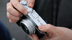 Hands holding a compact rangefinder camera with manual dials and a fixed lens.