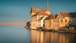 Yellow and cream-colored castle with multiple spires reflected in still blue water at golden hour.
