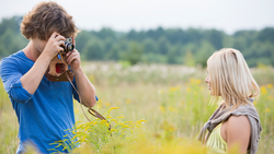 Young photographer with curly hair in blue shirt holding vintage camera up to face while photographing blonde woman in field.