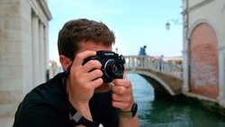 Photographer holding a Fujifilm medium format camera up to their face while standing in a European waterside alley.