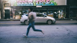 Blurred figure jogging across urban street past parked silver sedan and storefront with graffiti.