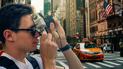 Photographer holding vintage film camera at street level, capturing urban cityscape with tall buildings and yellow taxi.