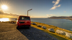 Red van parked on a coastal headland at sunset, overlooking a bay with mountains in the distance.