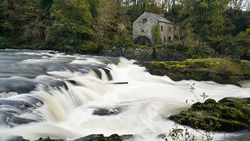 Cascading waterfall with stone mill building surrounded by trees on riverbank.