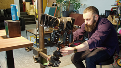Photographer adjusting a large format camera mounted on a wooden stand in a studio workspace.