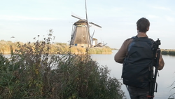 Photographer with camera and backpack standing on grassy bank overlooking traditional Dutch windmill across still water.