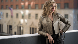 Woman with curly blonde hair leaning against a concrete railing overlooking urban buildings.
