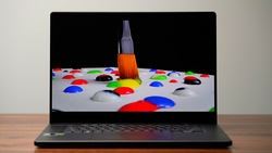 Screenshot of a paintbrush with orange bristles positioned above colorful paint droplets on a white surface displayed on a laptop screen.