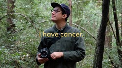 Person in forest holding vintage camera, wearing glasses and dark jacket.