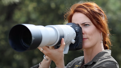 Photographer with reddish-brown hair holding a white telephoto lens to her eye while looking through the viewfinder.