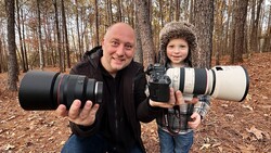 Adult and child holding professional camera lenses outdoors in a forest setting.