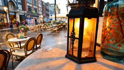 Illuminated black lantern on outdoor café table at dusk with blurred storefronts and diners in background.