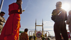 Buddhist monks in red and gold robes stand near a bridge with golden spires during a daytime ceremony.