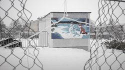 Winter mural viewed through chain-link fence showing snowman and figures in snowy landscape.
