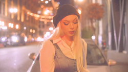 Woman with long blonde hair and dark beanie on urban street at night with warm city lights.
