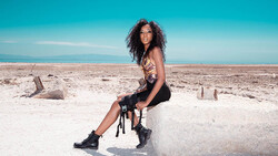 Woman posing on beach with clear sky, wearing patterned crop top and black shorts with boots.