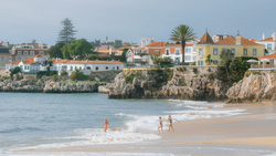 Swimmers in turquoise water below a clifftop coastal town with colorful buildings and sandy beach.