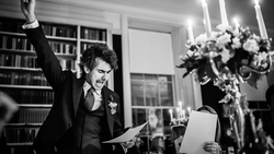 Groom in black tuxedo raising his fist triumphantly while holding papers in an elegant library setting.