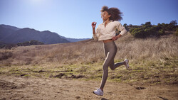 Woman jogging on an open trail through dry grassland with mountains in the distance under clear blue sky.